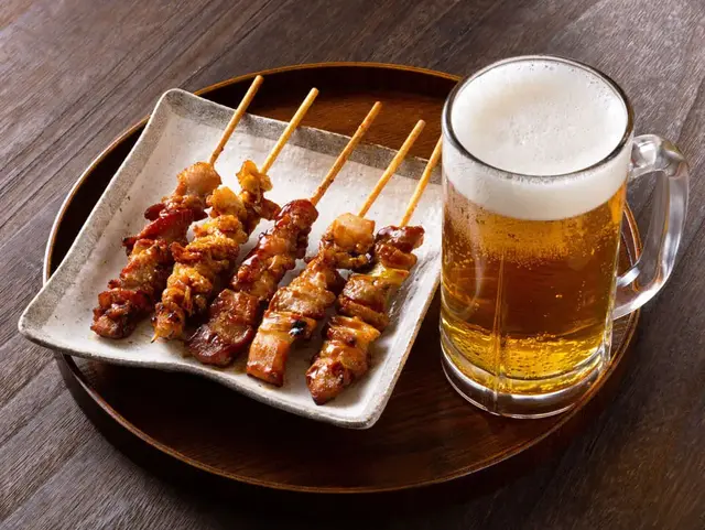 Yakitori (gegrillte Hähnchenspieße) auf Teller mit Bierglas daneben auf Holztisch.