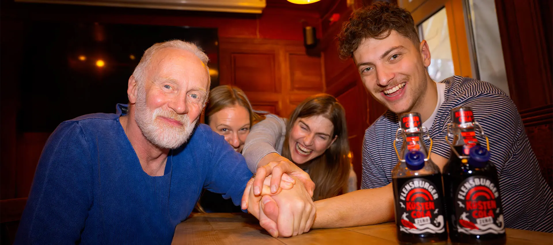 Gruppe von Leuten im Pub lachen, Hände übereinander auf dem Tisch, zwei Flensburger-Küstencola‑Flaschen im Vordergrund.