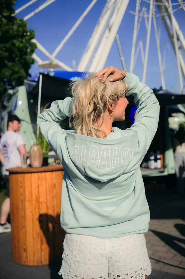 Junge Frau in mintfarbenem Hoodie mit ALOAHOI-Aufdruck, Rückenansicht, Kieler Woche und Riesenrad im Hintergrund.
