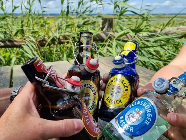 Es wird mit vier verschiedenen Flaschen angestoßen, zwei davon Braunglas, eine klar, eine blau, dahinter grüne Pflanzen und Wasser im Hintergrund.