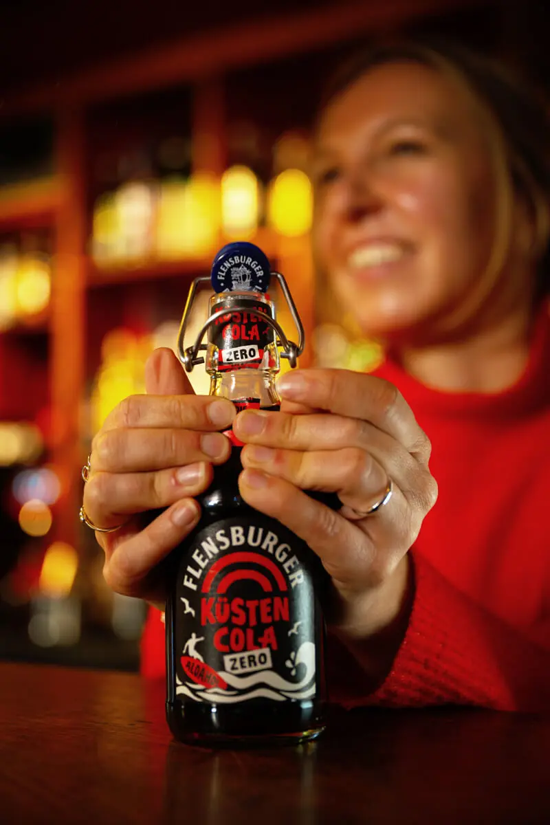 Frau im roten Pullover öffnet eine Flasche Flensburger Küstencola Zero in einer Bar.