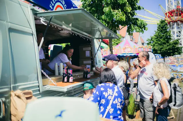 Küstenlimo-Getränke-Van auf der Kieler Woche 2025, Besucher bestellen Getränke, Bügelverschlussflaschen auf der Theke, bunte Fahrgeschäfte im Hintergrund.