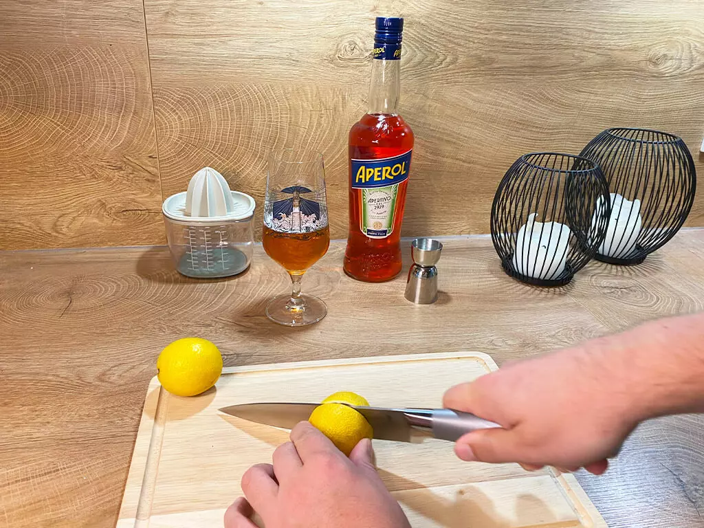 Eine Zitrone wird auf einem Holzbrett geschnitten, im Hintergrund eine Aperol-Flasche, Flensburger Edles Helles im Bierglas und eine Zitronenpresse.