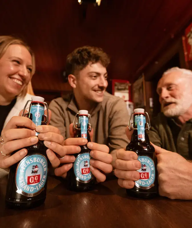 Drei Leute in Kneipe stoßen mit Flensburger Strand-Lager 0,0 % alkoholfreiem Bier an.