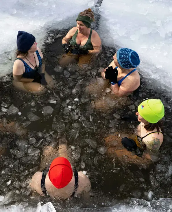 Gruppe beim Winterbaden in einem Eisloch, Menschen in Badeanzügen mit bunten Mützen.