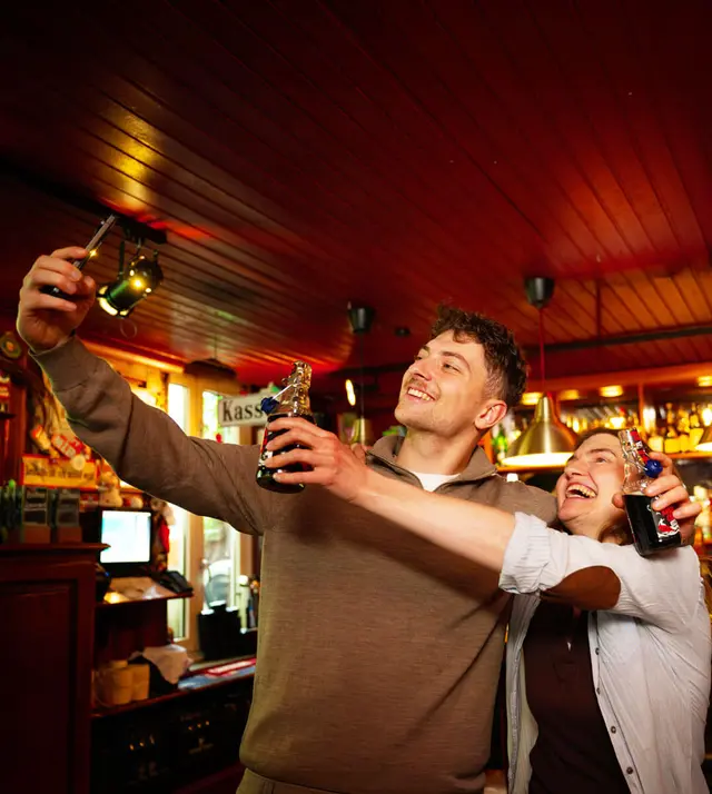 Zwei Freunde machen ein Selfie an der Bar, lachen und halten Bügelverschlussflaschen mit Küstencola Zero in gemütlicher, warm beleuchteter Kneipenatmosphäre.