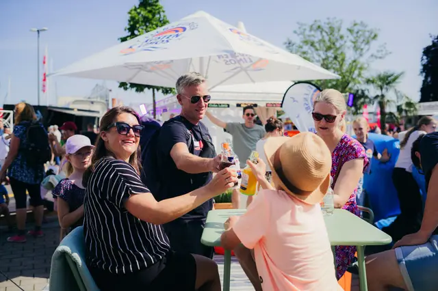 Familie und Freunde stoßen an einem Getränke-Tisch unter Sonnenschirm im Freien bei der Kieler Woche 2025 an.