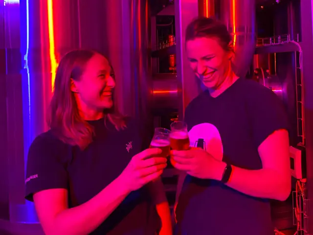 Die Brauerinnen Lisa & Karolin von FLENS stoßen vor der pink und blau beleuchteten Entalkoholisierungsanlage mit einer Bierprobe an.
