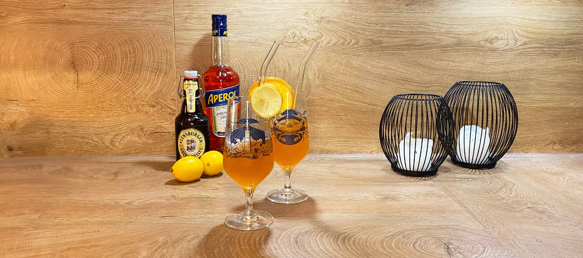 Zwei Gläser mit Aperol-Bier-Cocktail auf Holzuntergrund, im Hintergrund eine Flasche Aperol, Flensburger Edles Helles und zwei Zitronen.