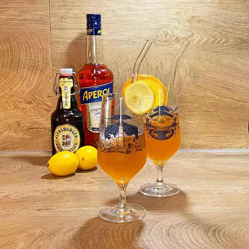 Zwei Gläser mit Aperol-Bier-Cocktail auf Holzuntergrund, im Hintergrund eine Flasche Aperol, Flensburger Edles Helles und zwei Zitronen.