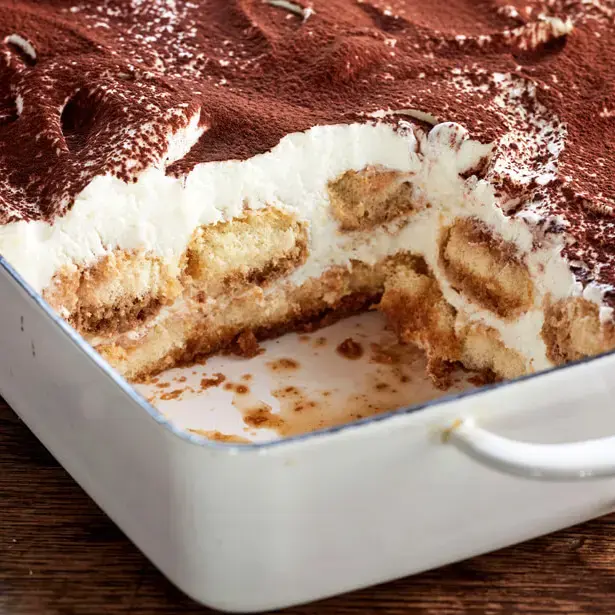Hausgemachtes Bieramisu (Tiramisu mit Bier) in Auflaufform mit Löffelbiskuits und Kakao auf rustikalem Holzbrett.