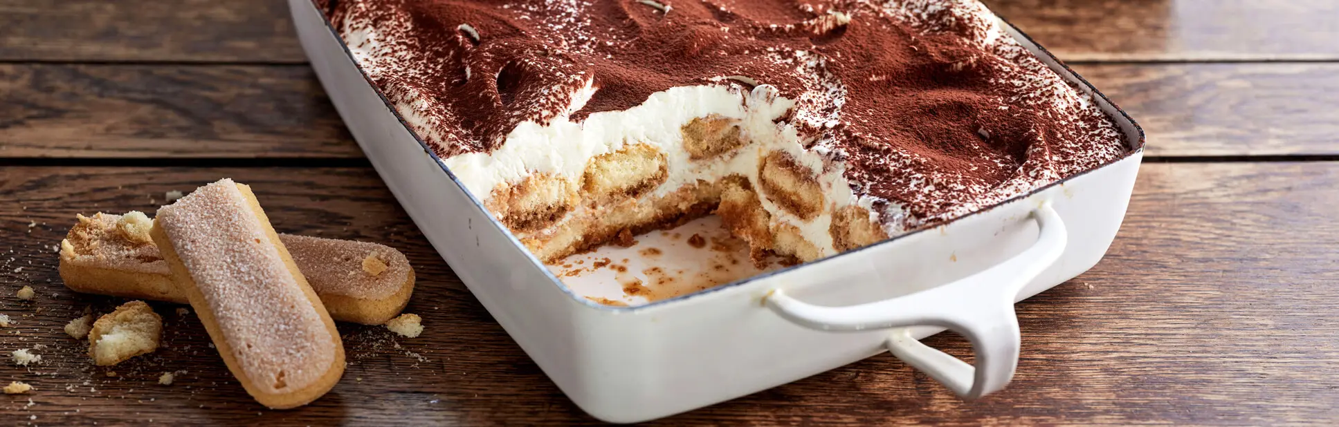 Hausgemachtes Bieramisu (Tiramisu mit Bier) in Auflaufform mit Löffelbiskuits und Kakao auf rustikalem Holzbrett.