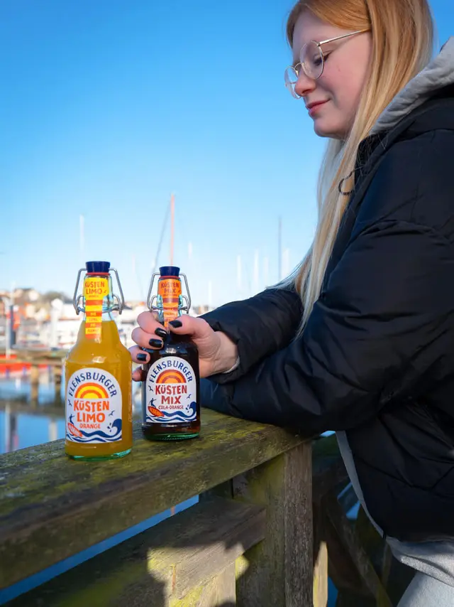 Eine Frau steht mit einer Küstenlimo in der Hand an einem Steg.