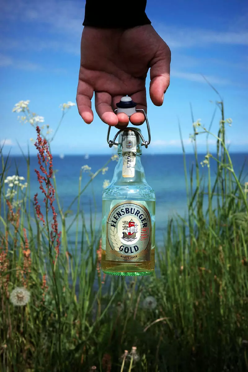 Eine Hand hält eine halb gefüllte Flasche Flensburger Gold auf einer Wiese vorm Wasser.