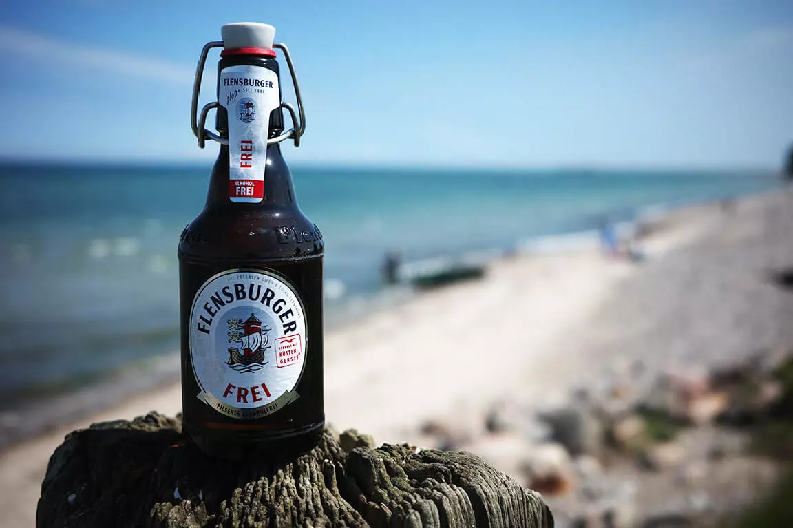 Eine Flasche Flensburger Frei auf einem Holzpoller am Strand.