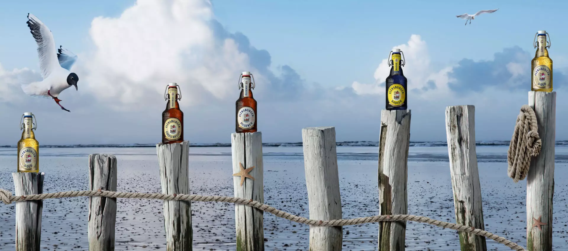 Vier Flensburger Flaschen stehen auf Holzstämmen am Meer.
