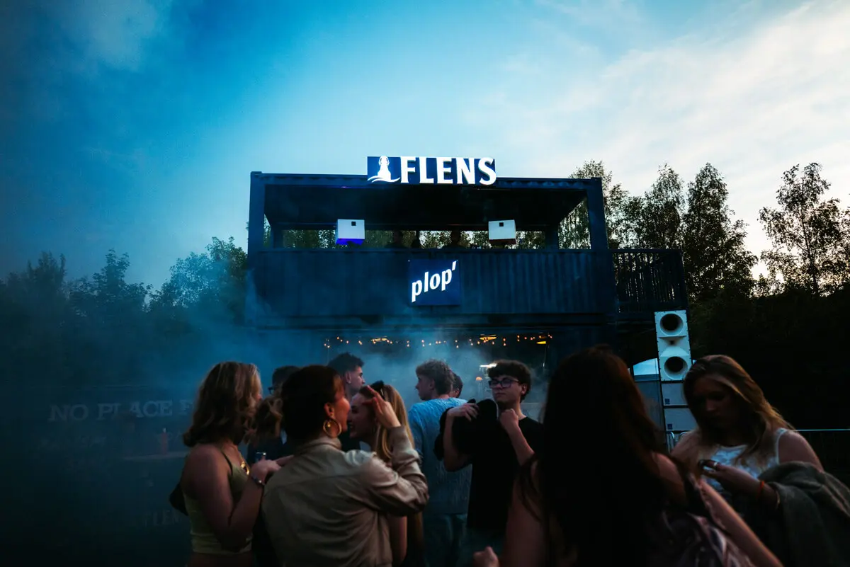 Butcher Jam 2026: Menschen vor Container-DJ-Stage mit Leuchtschrift 'FLENS' und 'plop' bei Abenddämmerung.
