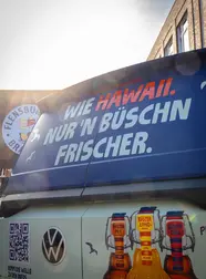 Der Küstenlimo-Bulli von hinten: Schriftzug "Wie Hawaii, nur 'n bischen frischer." auf blauem Hintergrund auf der Heckscheibe.