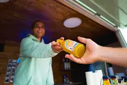 Eine Frau reicht eine Flensburger Küstenlimo Orange an einen Gast aus dem Küstenlimo-Promo-Van heraus.