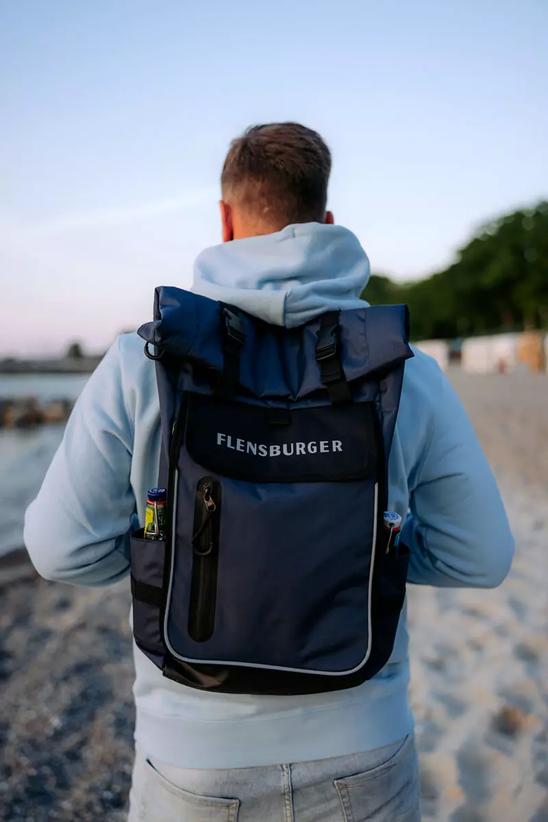 Ein Mann mit einem Rolltop-Rucksack in Blau und mit FLENSBURGER-Logo am Strand.