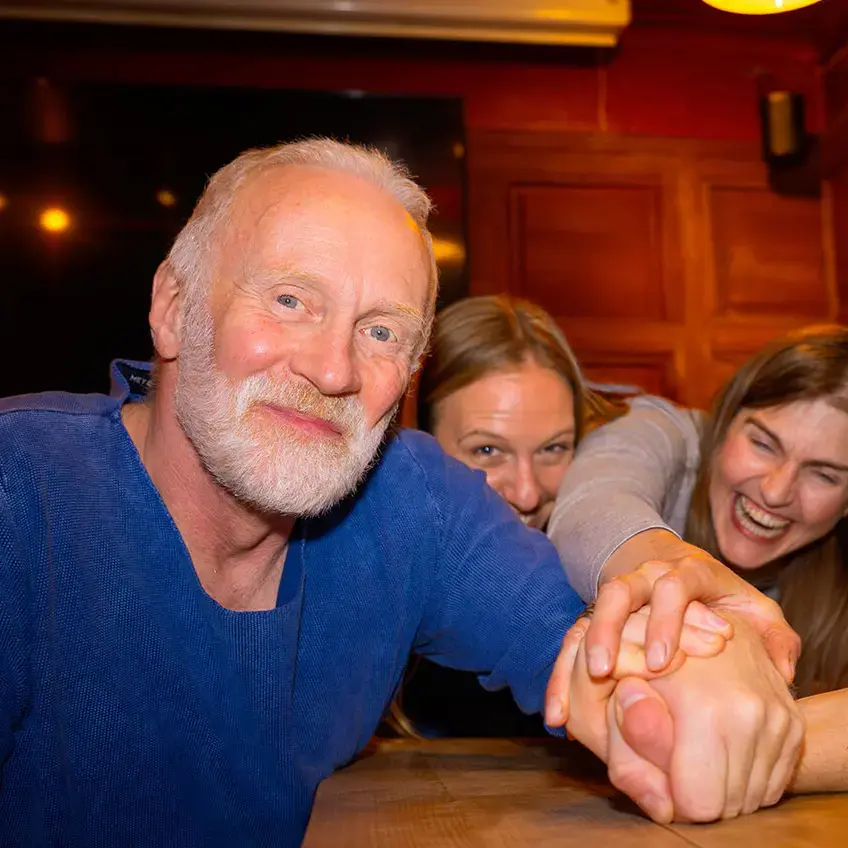 Gruppe von Leuten im Pub lachen, Hände übereinander auf dem Tisch, zwei Flensburger-Küstencola‑Flaschen im Vordergrund.