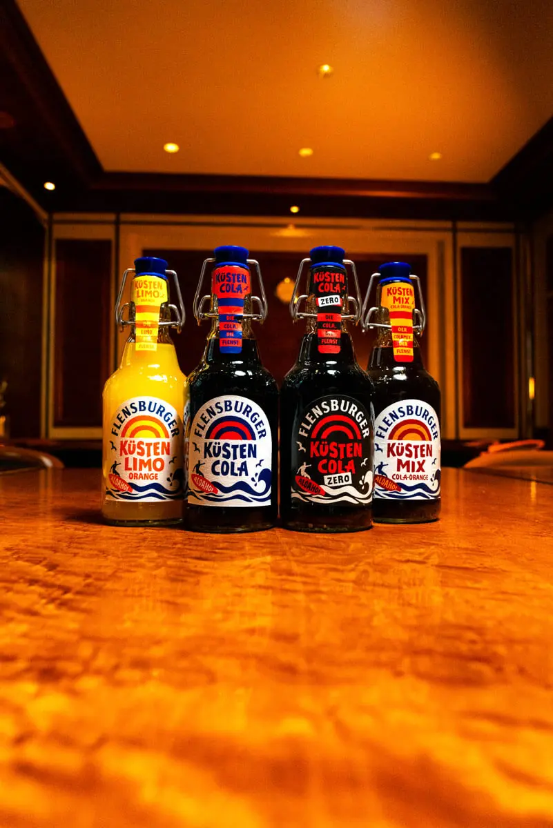 Vier Flensburger Küstenlimos in Glasflaschen auf Holztisch, warmes Restaurantambiente.