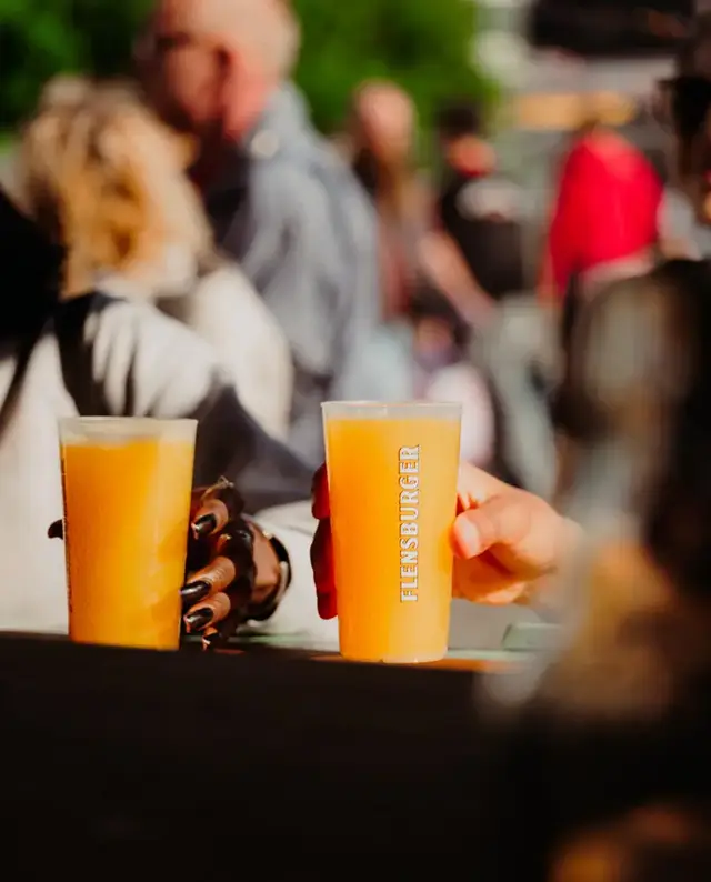 Zwei Hände halten Flensburger-Becher mit Flensburger Küstenlimo Orange vor Menschenmenge auf Outdoor-Event.