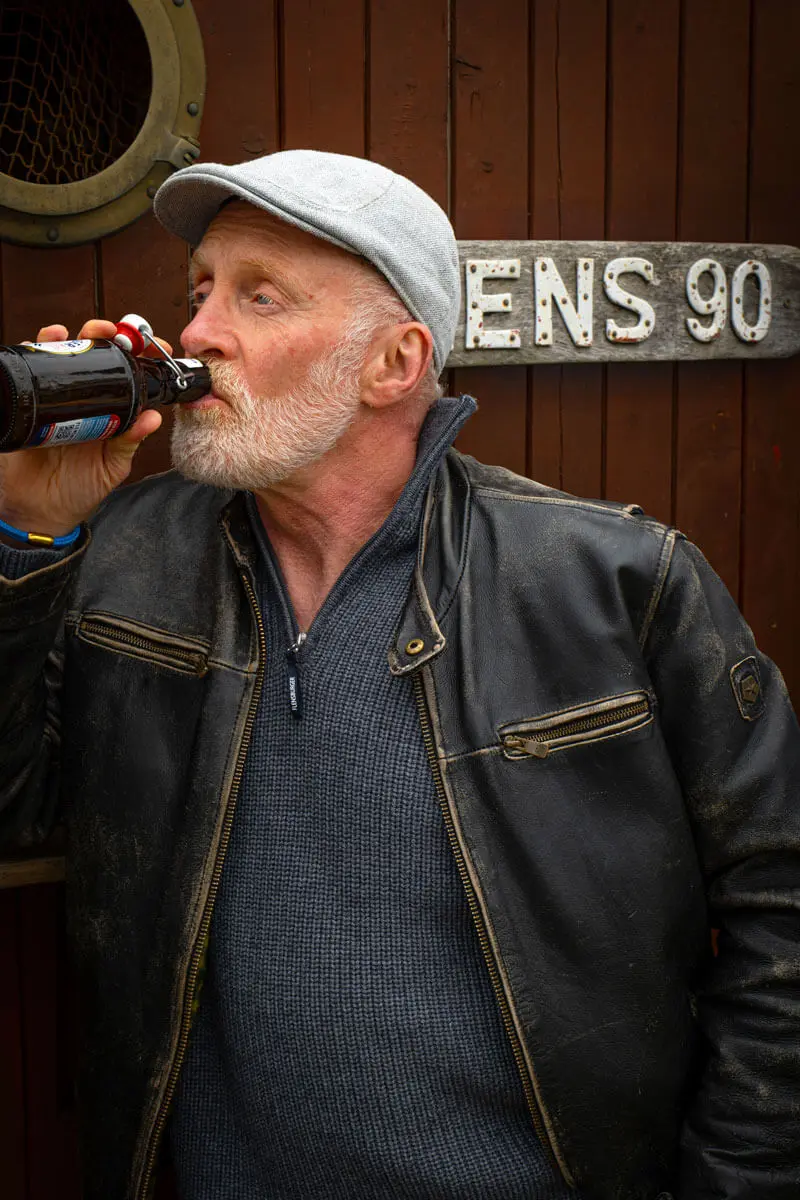 Andreas Arndt in Lederjacke und Schirmmütze trinkt FLENS Bier aus Bügelflasche vor Holztür.