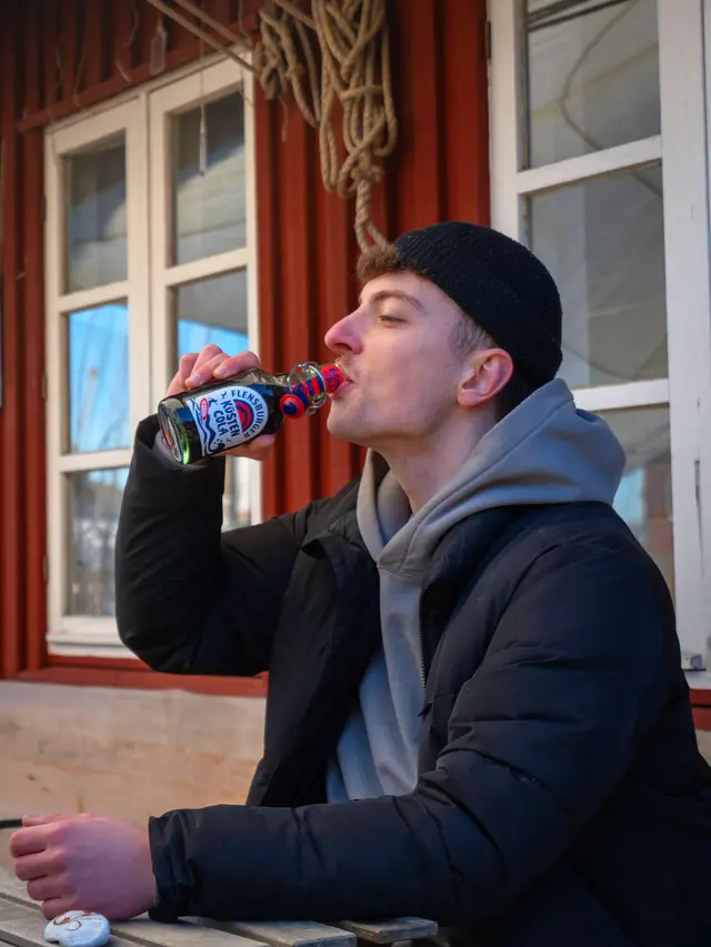 Junger Mann trinkt Flensburger Küstencola aus Flasche vor rotem Bootshaus.