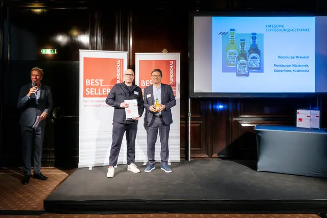 Preisverleihung für den RUNDSCHAU Bestseller Award 2025: Zwei Personen mit Urkunde und Küstenlimo Orange auf Bühne vor Bestseller-Banner und Produktpräsentation.
