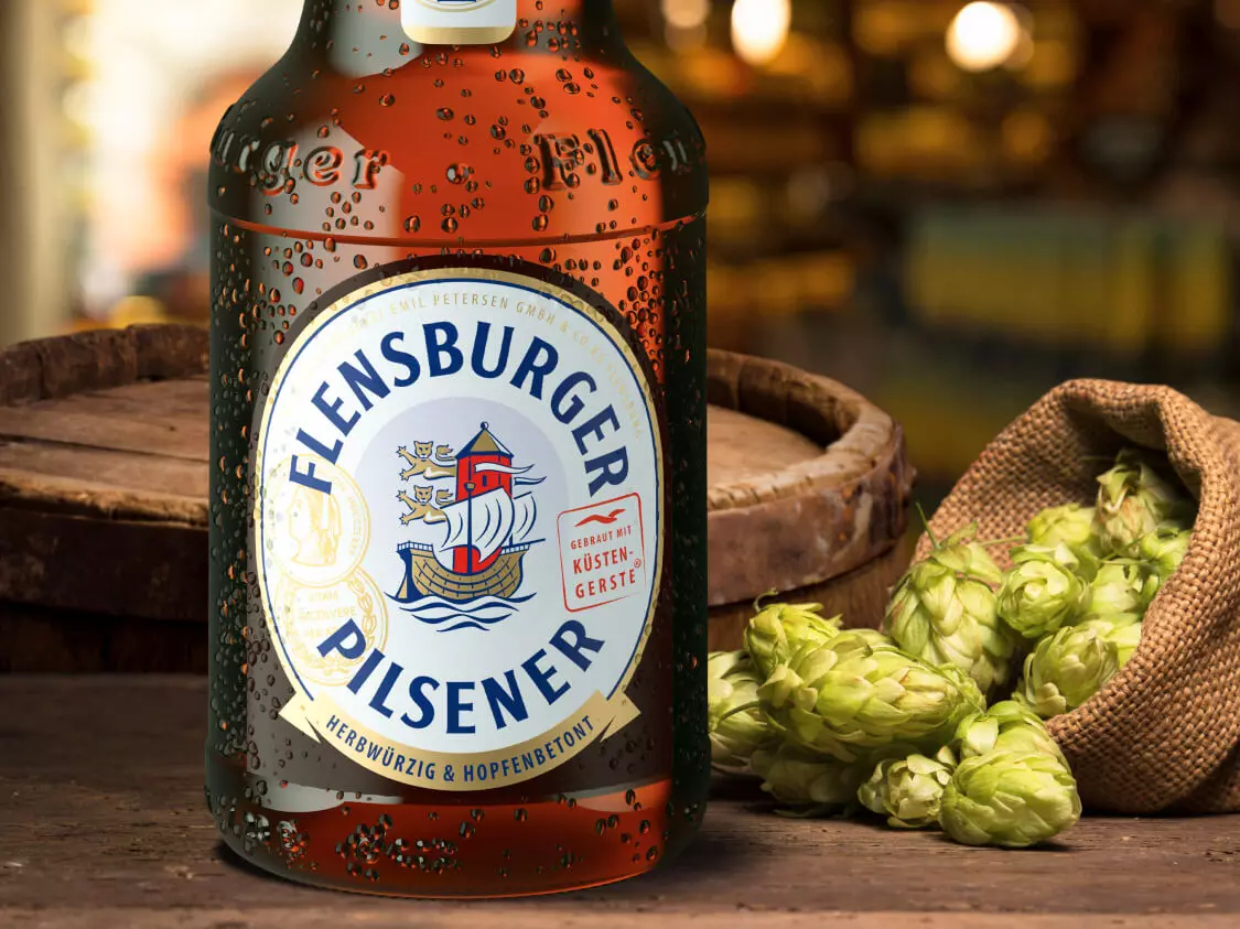 Hopfendolden in einem Jutesack auf einem Tisch, daneben eine Flasche Flensburger Pilsener.