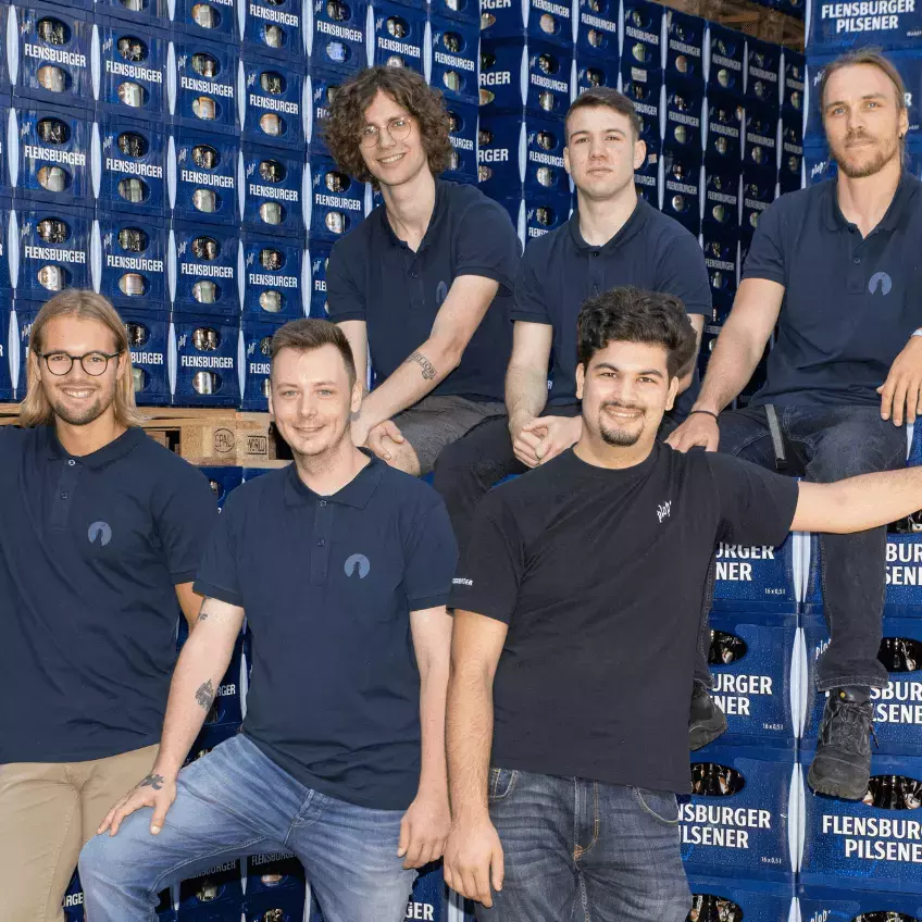 Fünf Flensburger Mitarbeiter, in blauen FLENS-Shirts vor Flensburger Kisten.