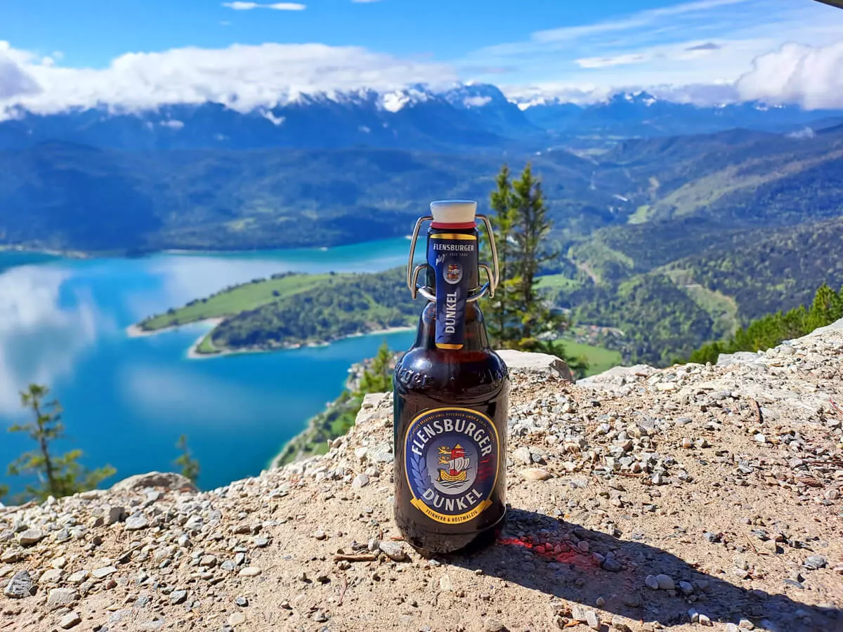Eine Flasche Flensburger Dunkel steht an einer Klippe vor einem sonnigen See- und Bergpanorama.