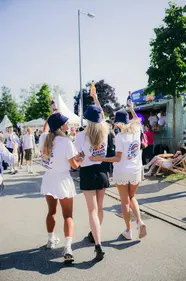 Drei junge Frauen bei der Kieler Woche 2025, Rückenansicht, halten Küstenlimoflaschen hoch, tragen weiße Shirts, Shorts und blaue Küstenlimo-Anglerhüte.