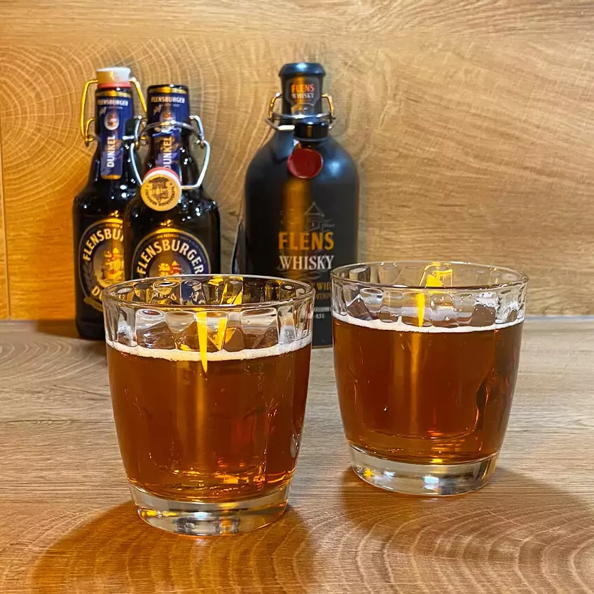 Zwei Tumbler-Gläser mit dem Boilermaker-Cocktail, bestehend aus Flensburger Dunkel und FLENS Whisky, mit den dazugehörigen Flaschen im Hintergrund.