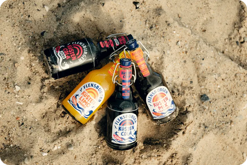 Flensburger Küstenlimo Orange, Küstencola, Küstencola Zero und Küstenmix im Sand am Strand.