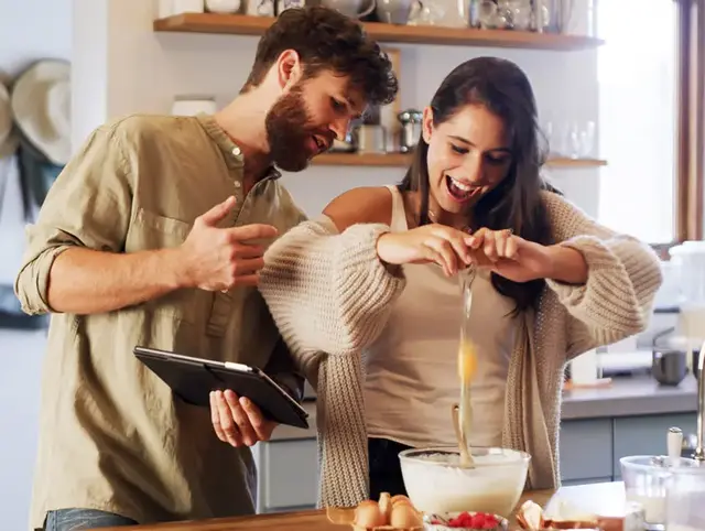 Paar bereitet Bieramisu (Tiramisu mit Bier) vor – Frau schlägt Ei in Schüssel, Mann hält Tablet mit Rezept.