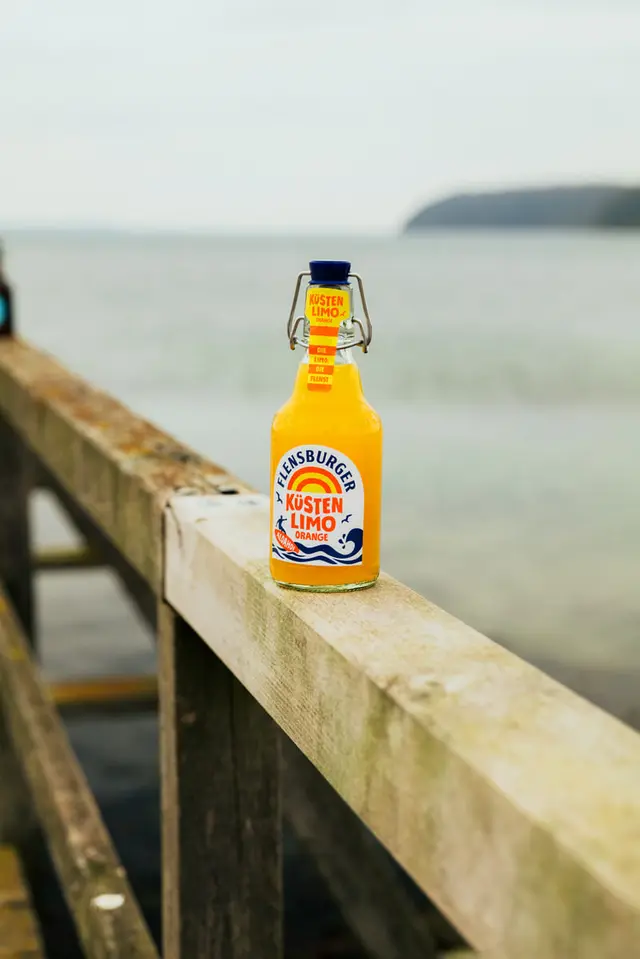 Flensburger Küstenlimo Orange auf dem Geländer eines Holzstegs am Meer.