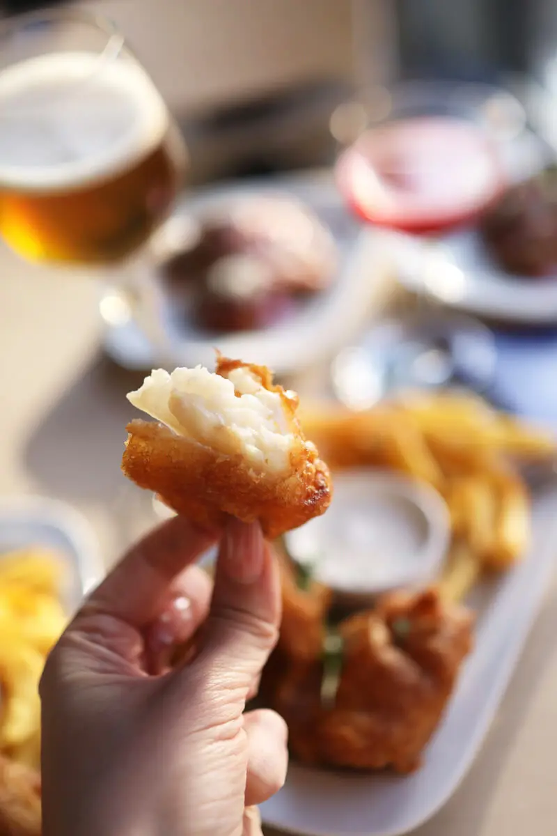 Hand hält knuspriges Stück Backfisch mit Pommes und Dip und Bierglas im Hintergrund.