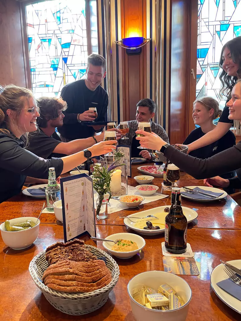 Gesellschaft, die mit Bier Flensburger Salon anstößt; auf dem Tisch einige Snacks.