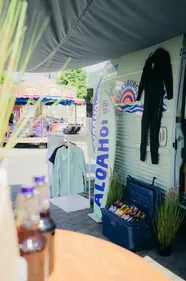 Aufbau am Küstenlimo-Van mit ALOAHOI-Surf-Outfits, Neoprenanzug, Deko-Surfboard und Getränkekühlbox.