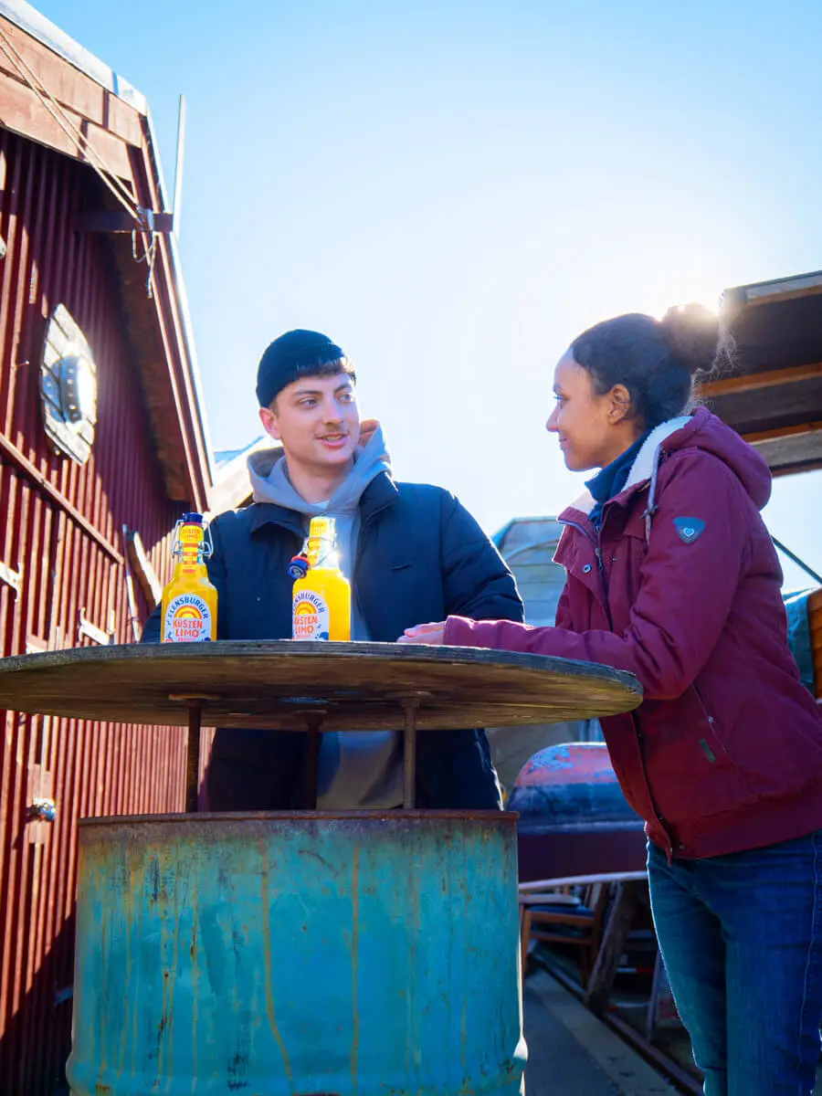 Zwei Personen an rustikalem Fass-Tisch mit Küstenlimo Orange vor rotem Holzhaus an sonnigem Tag.