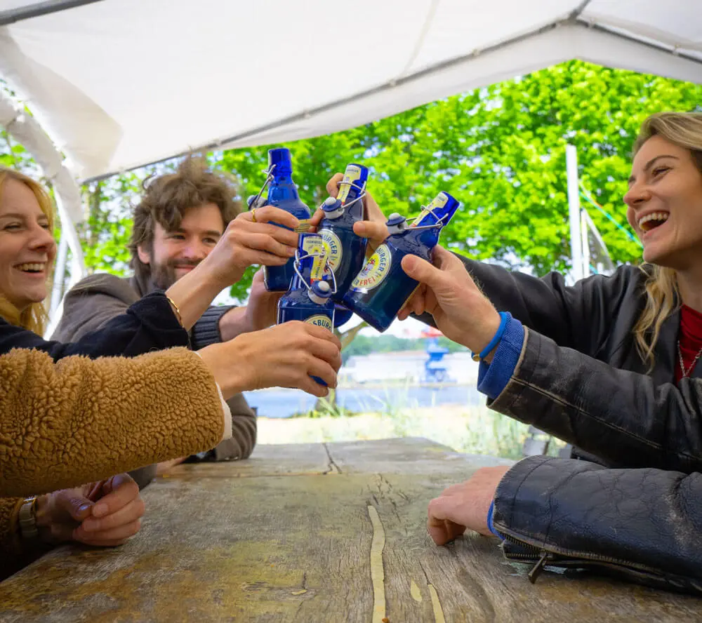 Menschen stoßen mit blauen FLENS Radler Frei in Bügelverschlussflaschen auf rustikalem Holztisch unter Zeltdach an.