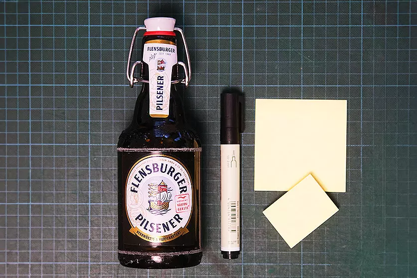 Auf einer grünen Bastelmatte liegen eine Flasche Flensburger Pilsener, ein schwarzer Permanentmarker und gelbe Haftnotizzettel.