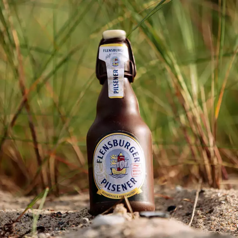 Flensburger Pilsener als Schokoladenflasche vor Dünengras im Sand.