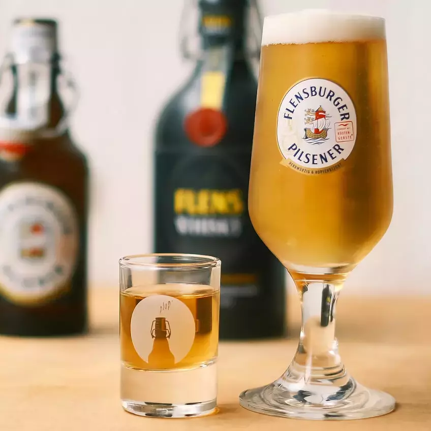 Ein Glas frisch gezapftes Flensburger Pilsener, daneben ein Shotglas mit FLENS Whisky; im Hintergrund etwas verschwommen die dazugehörigen Flaschen.