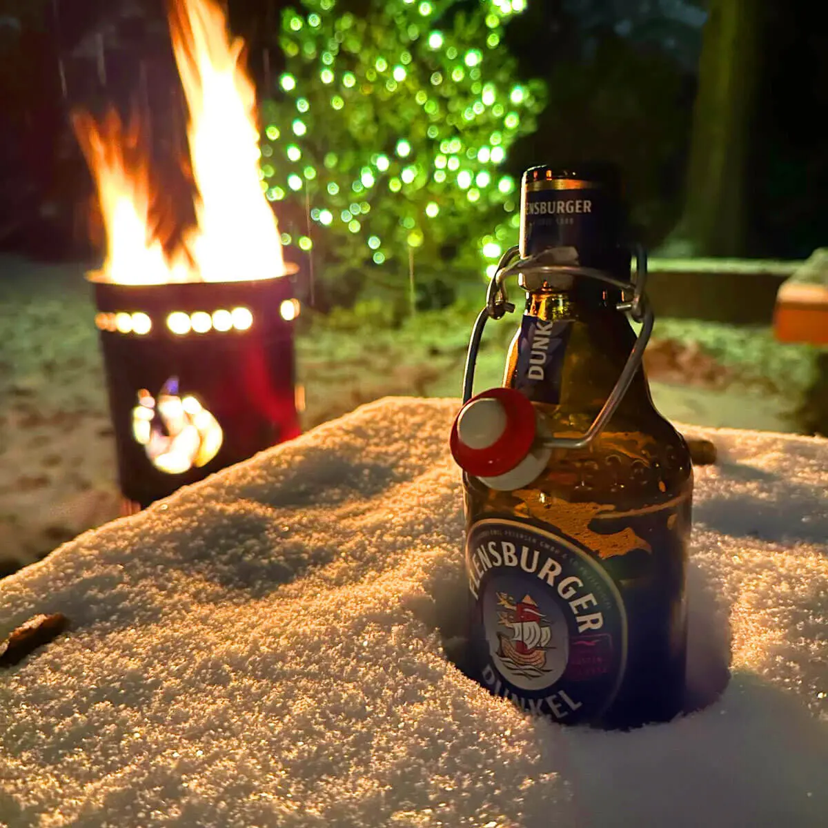 Flensburger Dunkel Bierflasche im Schnee neben Lagerfeuer und Lichterketten bei Nacht