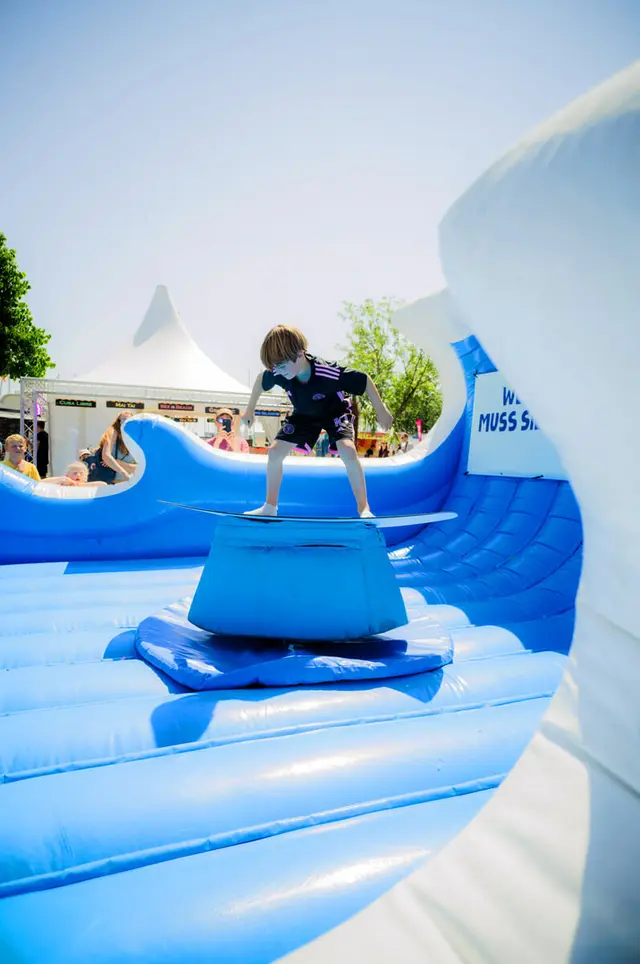 Junge surft auf einem blauen aufblasbaren Bullriding-Surfbrett bei der Kieler Woche 2025 am Küstenlimo-Stand.