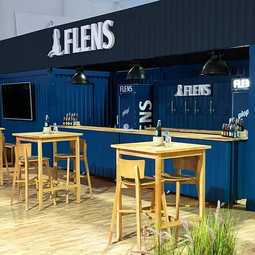 FLENS Messestand im blauen Container-Look mit Holztischen, Barhockern und Flensburger Bierflaschen.