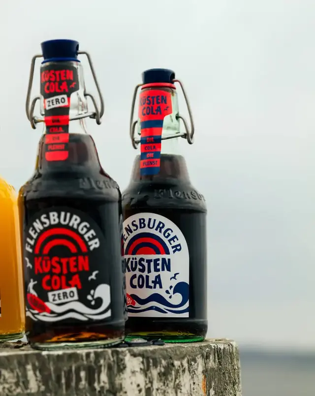 Zwei Flaschen Küstencola von Flensburger mit Bügelverschluss auf verwittertem Pfahl vor Meereshorizont.